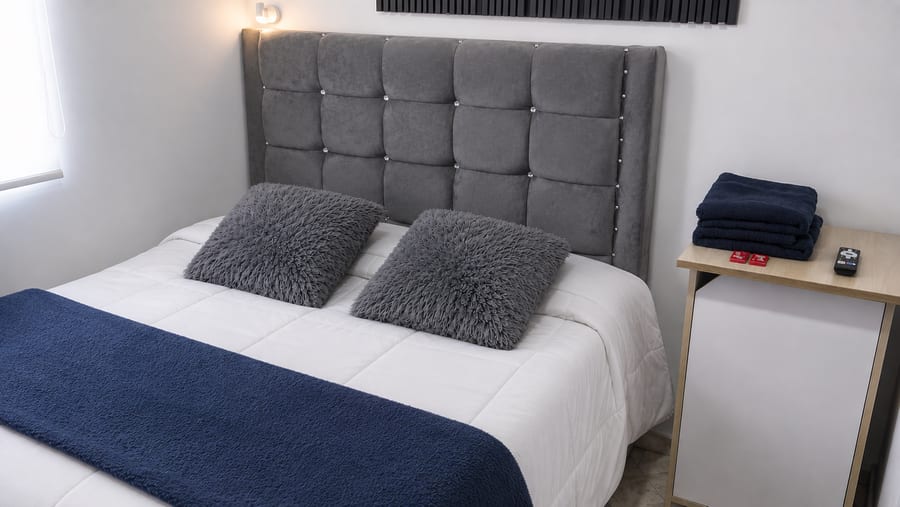 Cama doble de la habitación 201 con cabecero gris capitone, ropa de cama blanca y pie de cama azul