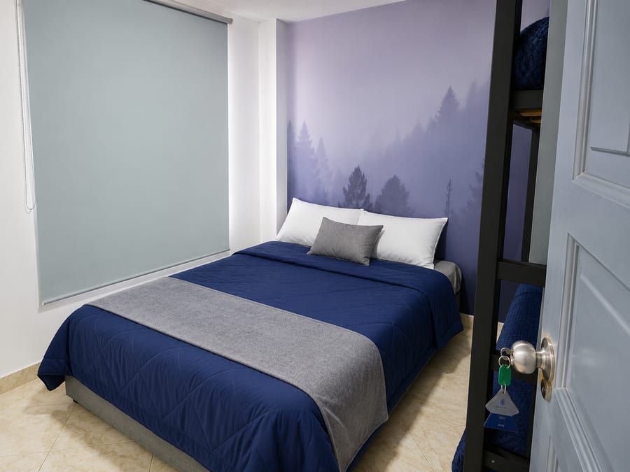 Cama doble de la habitación 202 con ropa de cama azul y gris, frente al mural de bosque