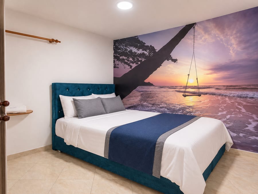 Habitación 203 con cama de cabecero azul capitone y mural de atardecer en la playa